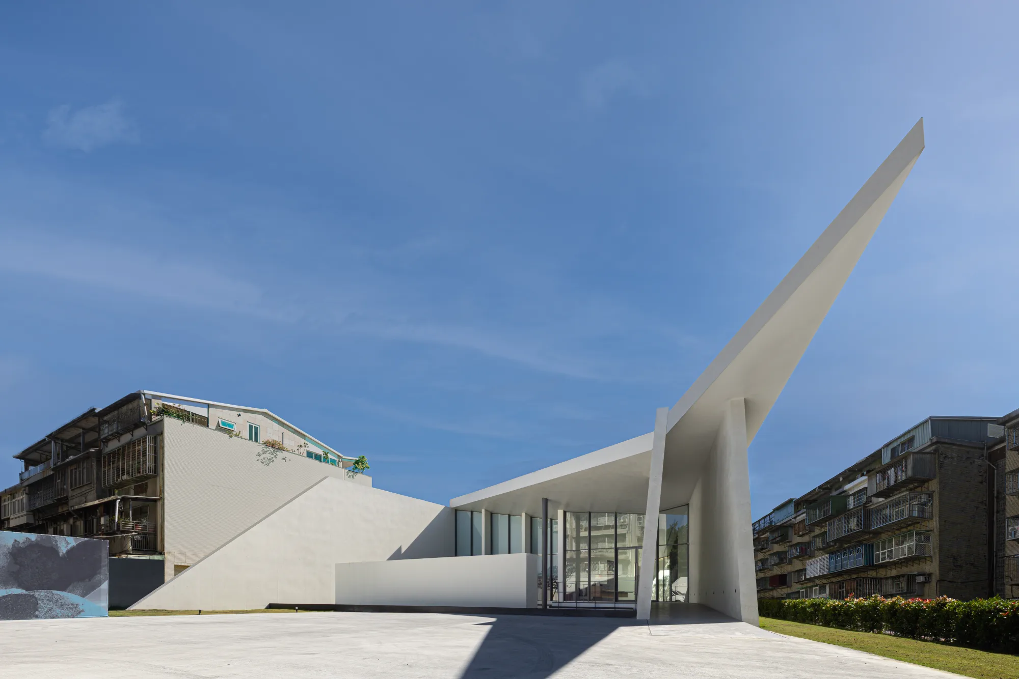 帛唐空間美學｜展售中心暨樣品屋 — 建築日間外觀，斜插天際的白色翼板與相互交切的量體面，清水混凝土表面在藍天下顯出張力，座落於台北士林的老社區地塊｜空間攝影 / 琢磨影像