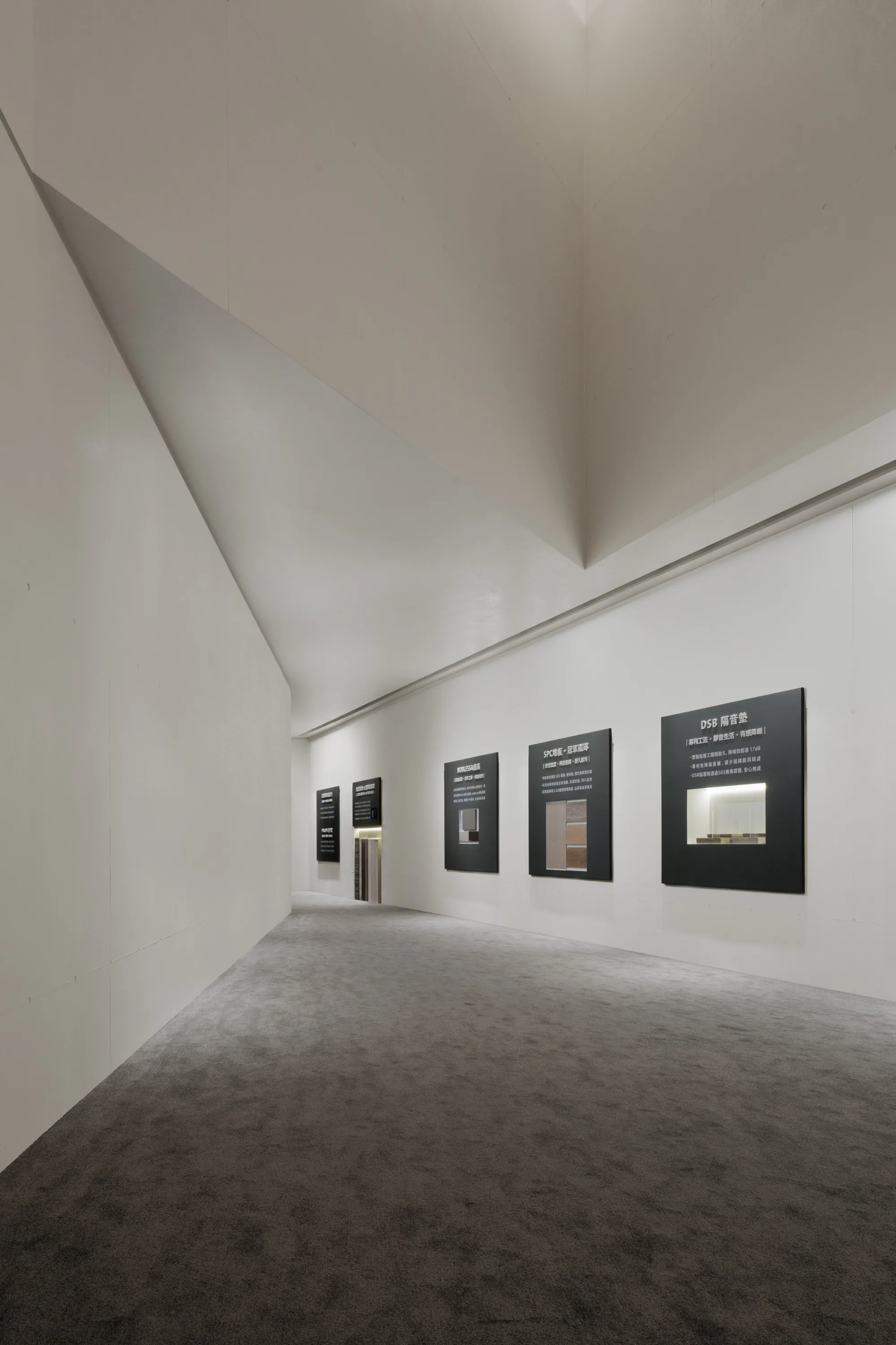 帛唐空間美學｜展售中心暨樣品屋 — 室內展示廊道，白色斜面天花斜落，牆面懸掛計畫展板，柔光鋪陳出安靜的觀展動線｜空間攝影 / 琢磨影像