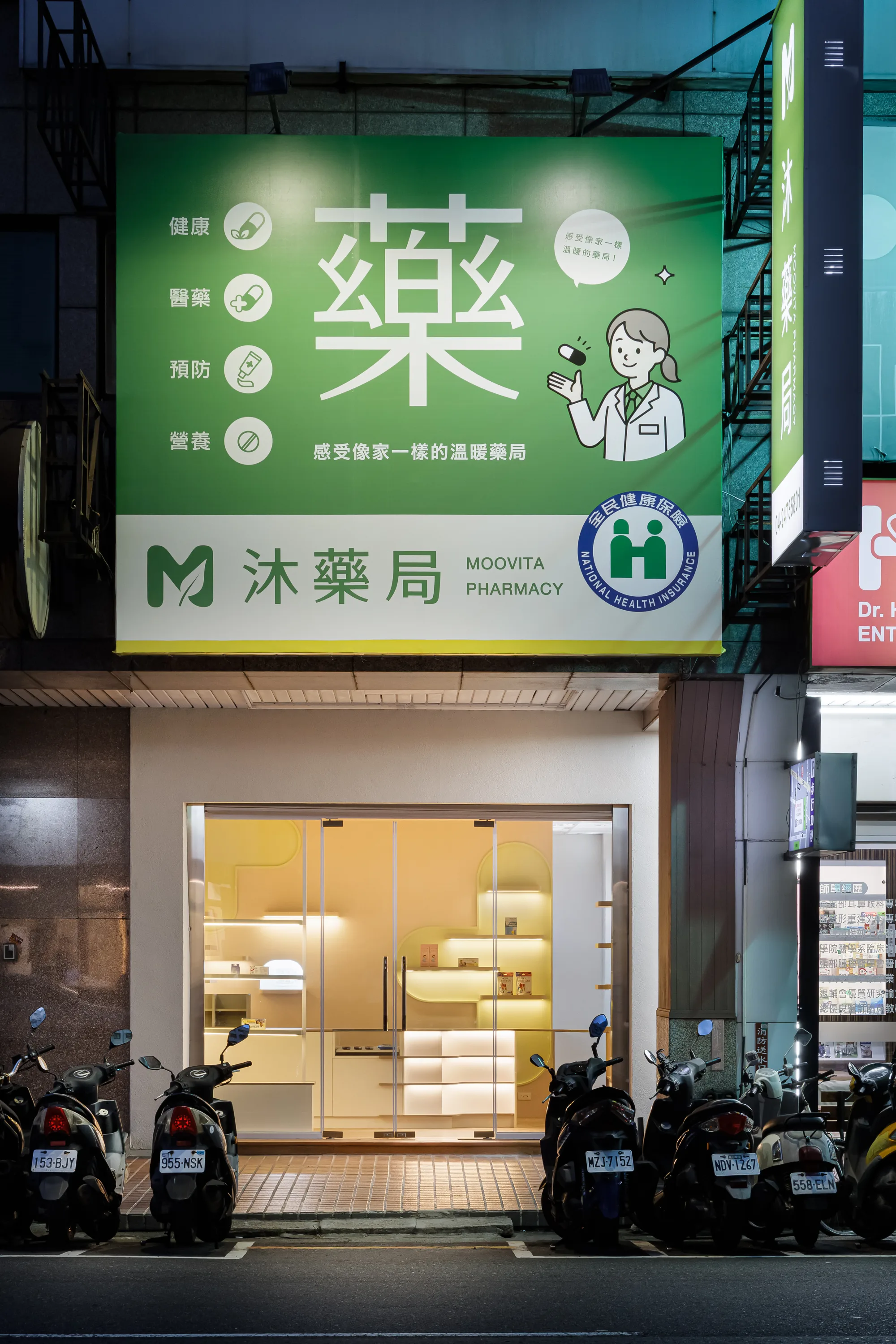 映墨設計|沐藥局 — 夜間店招,綠色『藥』字燈箱與『沐藥局』招牌點亮街邊店面,溫暖室內光透出玻璃門面,形塑親切的社區藥局意象|空間攝影 / 琢磨影像