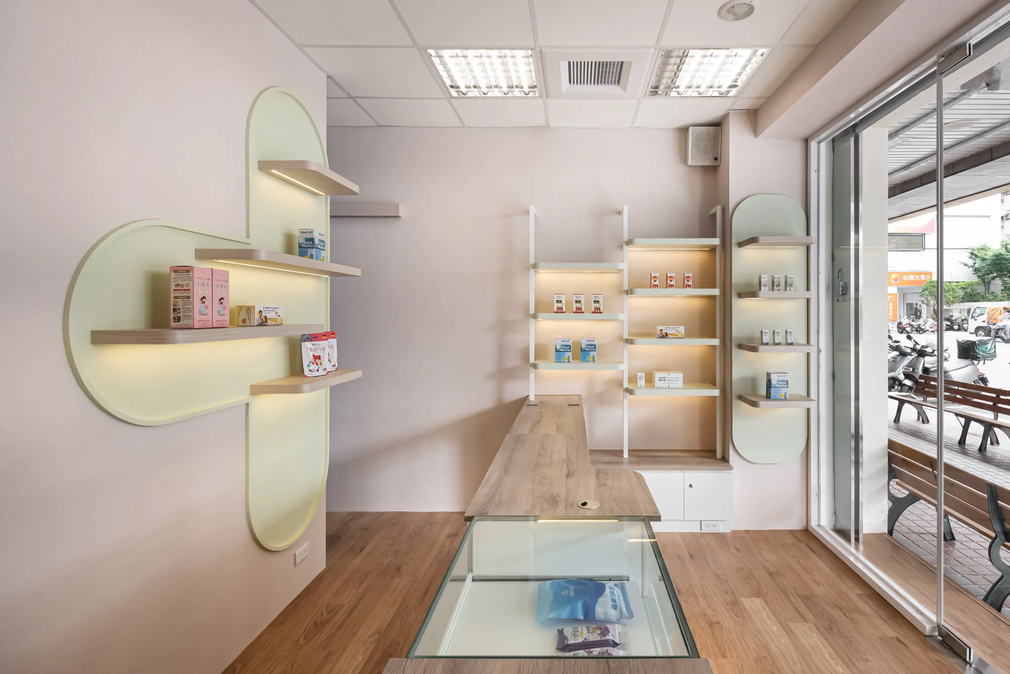 映墨設計｜沐藥局 — 店內全景，柔粉牆面與薄荷綠拱形造型牆結合木質浮層板與暖色間照，臨街整面落地玻璃引入日光，地面嵌入玻璃展示櫃與淺木地坪，鋪陳療癒系藥局空間｜空間攝影 / 琢磨影像