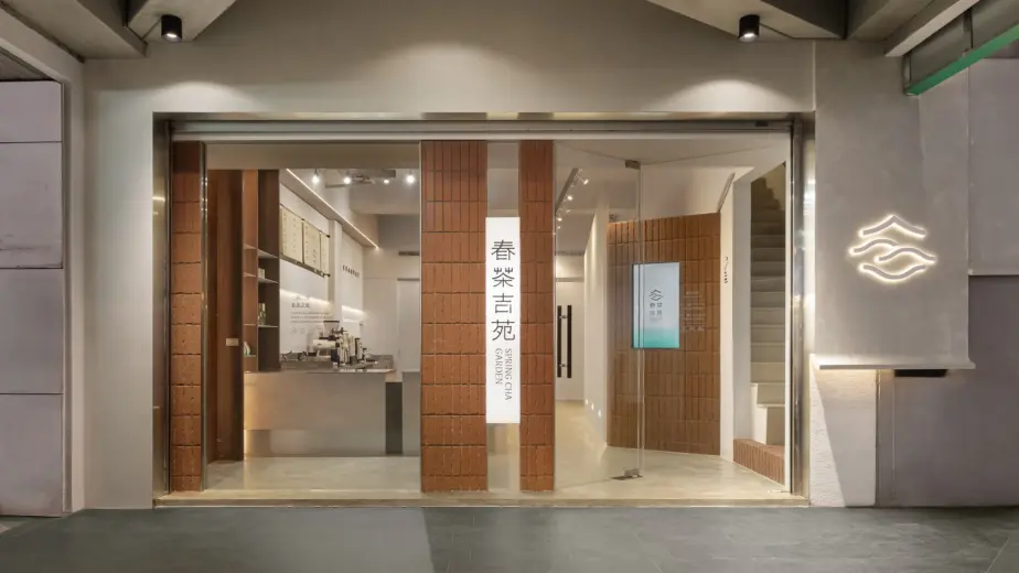 春茶吉苑 — 入口門面,清水模門廊與紅磚立柱框出垂直招牌布幔「春茶吉苑 SPRING CHA GARDEN」,品牌標誌燈點亮兩側,內部吧台隱約可見,塑造沉靜雅致的茶飲品牌入口｜空間攝影 / 琢磨影像